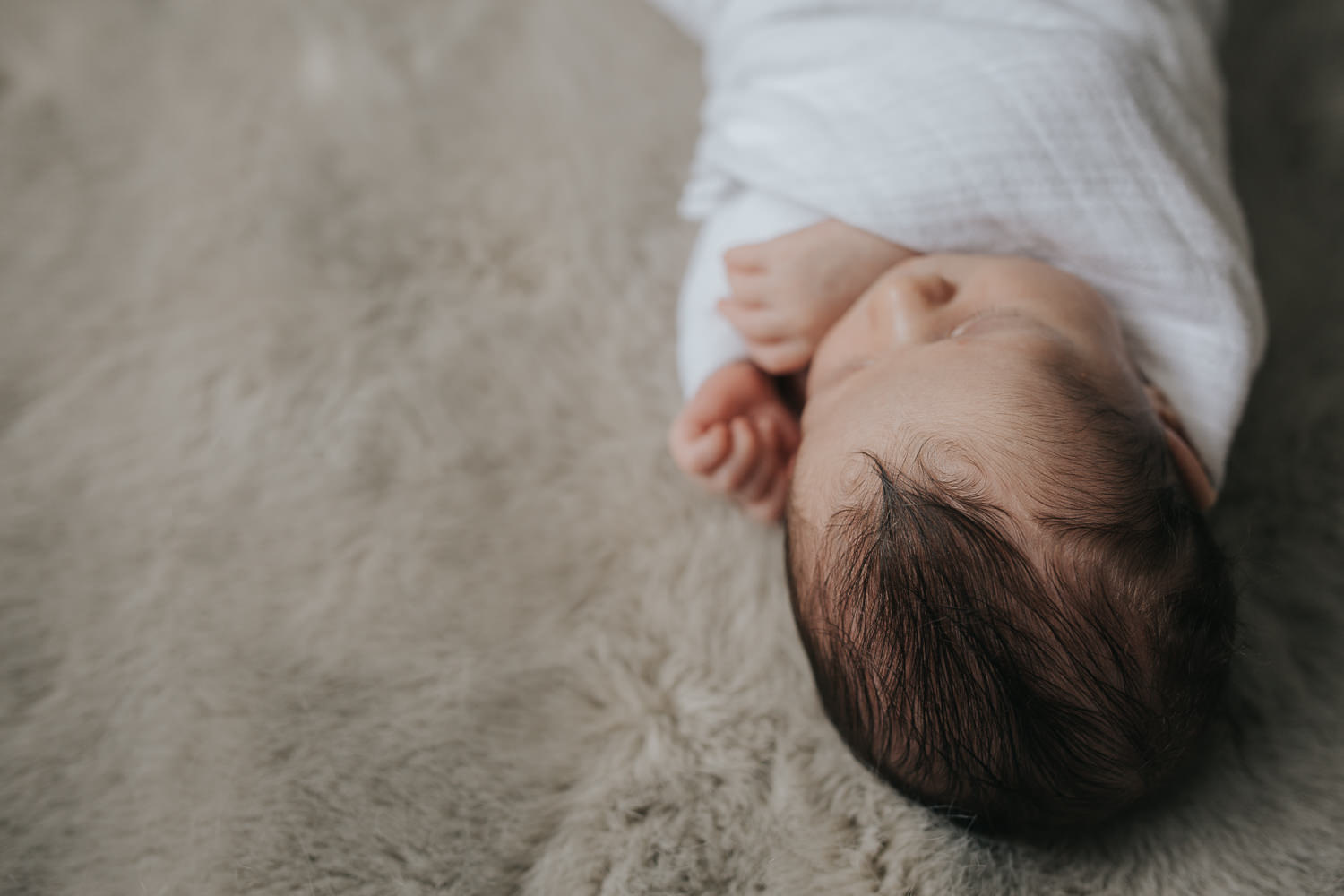 2 week old baby boy with dark hair lying on fur blanket in white swaddle, sleeping with hands next to face - GTA Lifestyle Photography