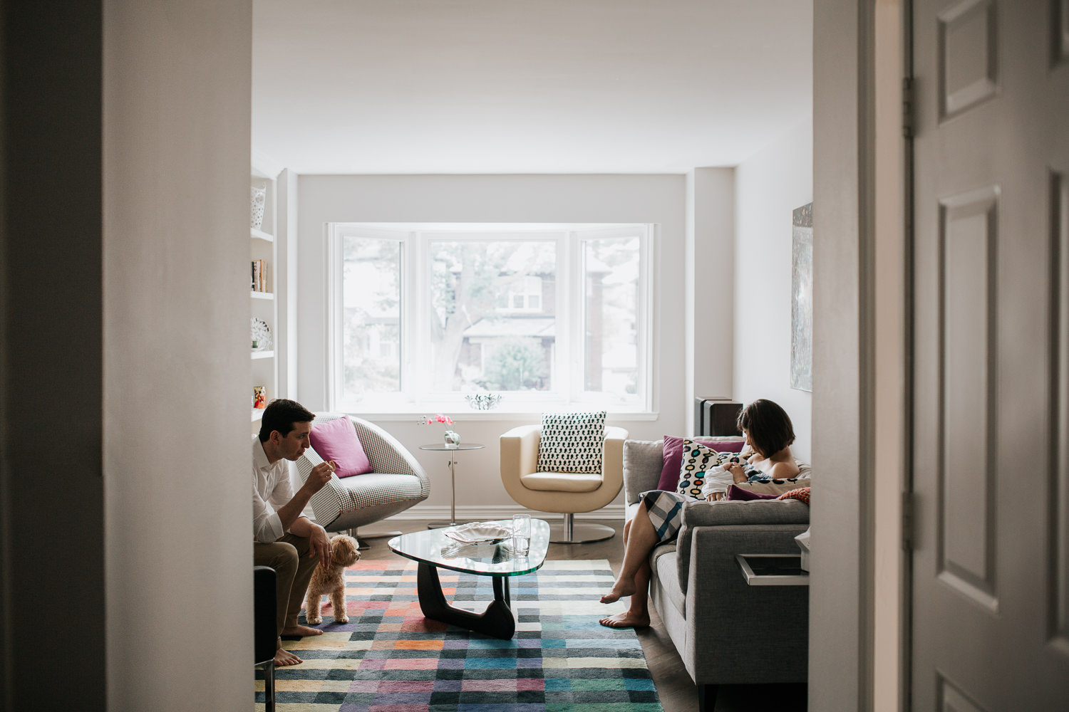 new mom with short brunette hair sitting on couch in living room snuggling 2 week old baby boy in white swaddle, dad and dog sitting across from them - Stouffville In-Home Photography