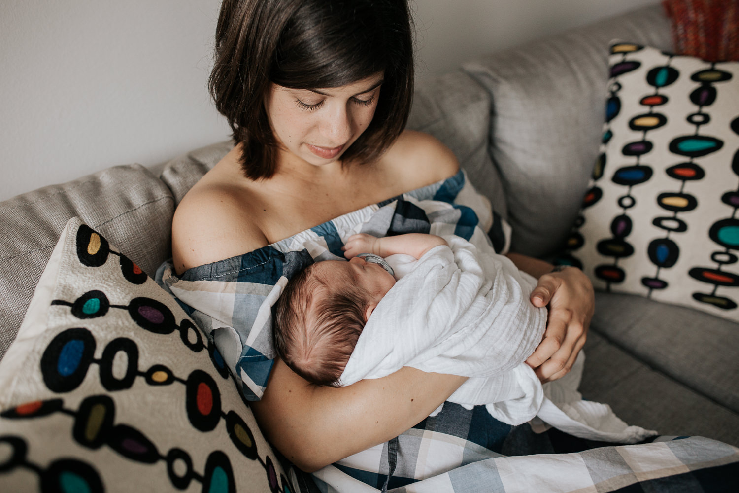 new mom with short brunette hair sitting on couch in living room snuggling 2 week old baby boy in white swaddle - Markham In-Home Photography