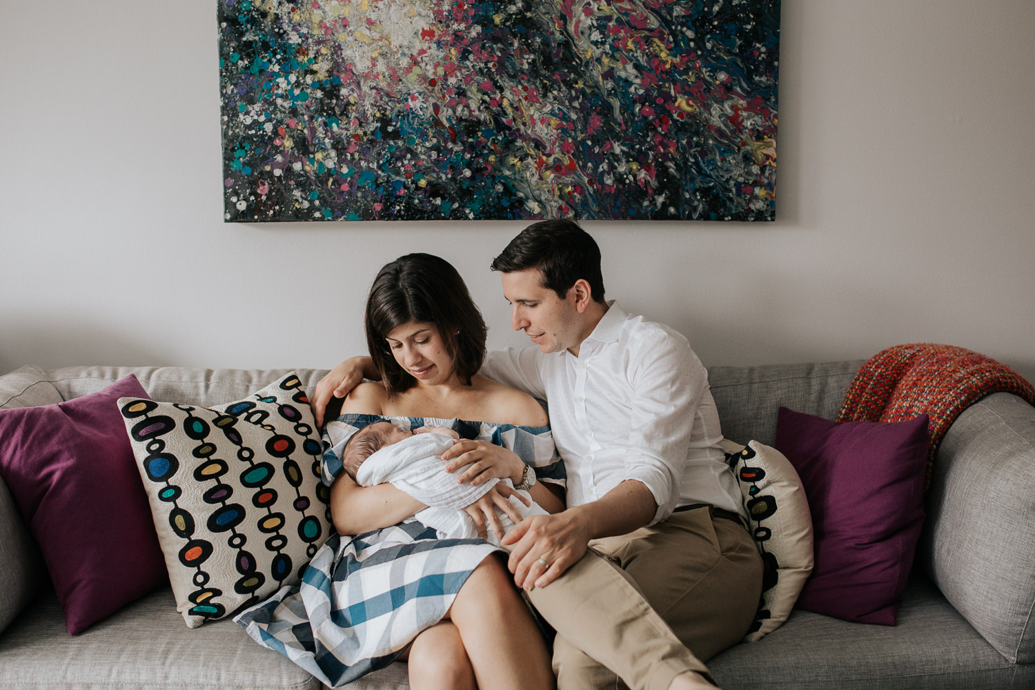 new parents sitting on couch in living room snuggling 2 week old baby boy in white swaddle, mom holding son, dad with arm around her - Barrie In-Home Photography