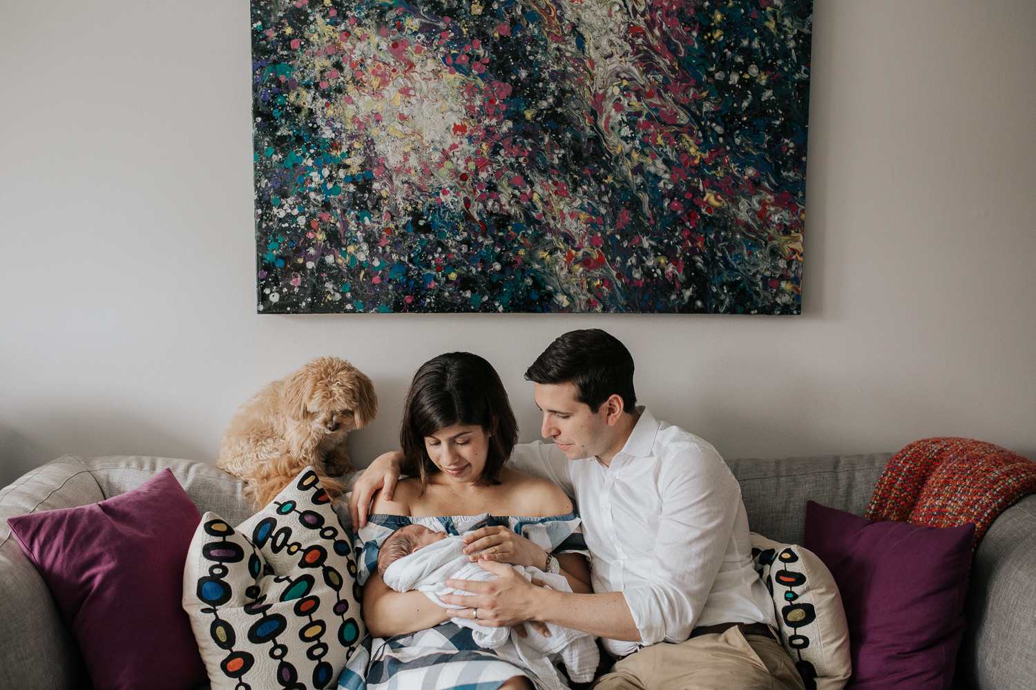 new parents sitting on couch in living room snuggling 2 week old baby boy in white swaddle, mom holding son, dad with arm around her and golden dog sitting on back of couch - GTA In-Home Photography