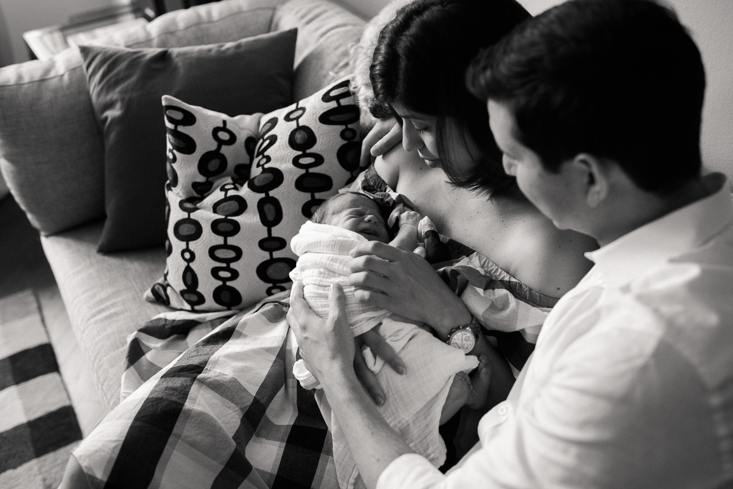 new parents sitting on couch in living room snuggling 2 week old baby boy in white swaddle, mom holding son, dad with arm around her - Newmarket In-Home Photos