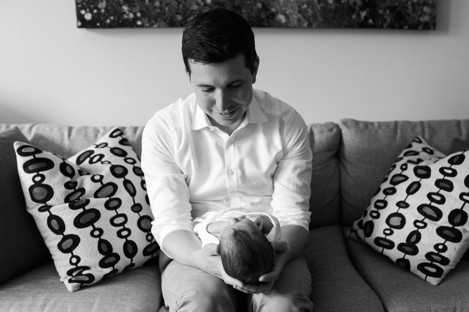 new father sitting on couch holding 2 week old baby boy in white swaddle, son's head resting in dad's hands - Barrie In-Home Photos