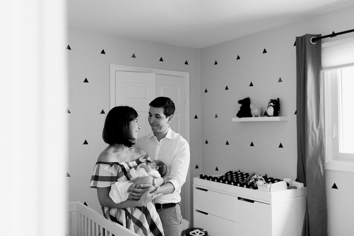 new parents standing in nursery, mom holding swaddled 2 week old baby boy with dark hair, dad looking at mom -  Newmarket In-Home Photography