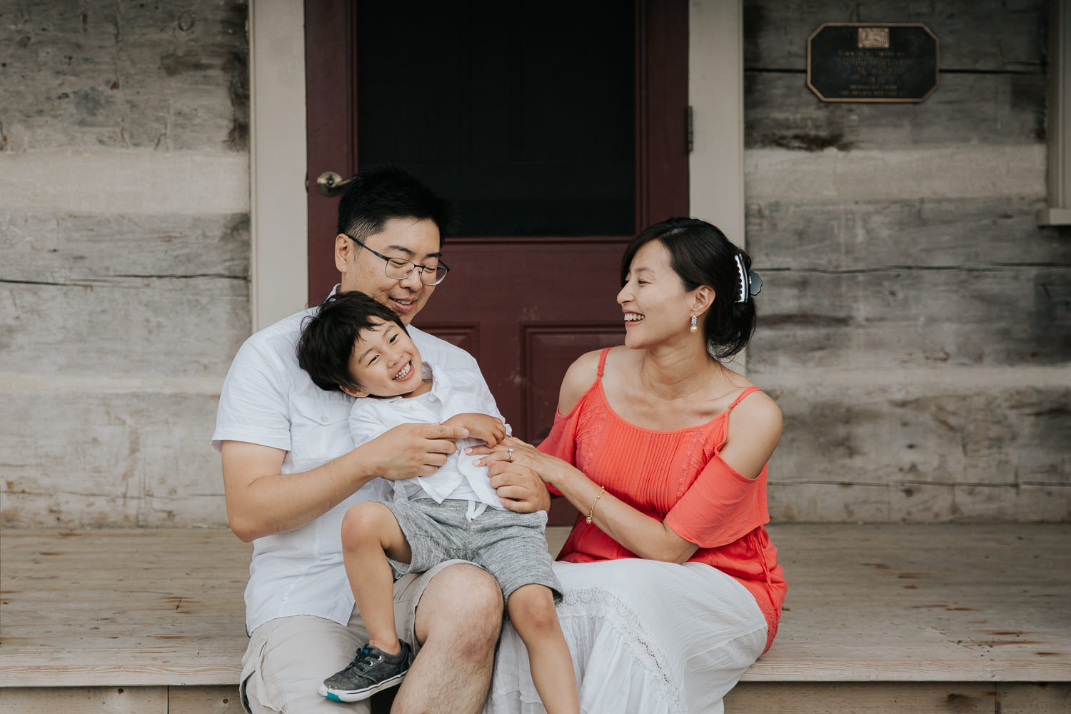 family of 3 sitting on front porch, toddler son sitting on dad's lap, laughing as mom tickles him, father and son in white shirts, mother wearing coral top - Newmarket In-Home Photography