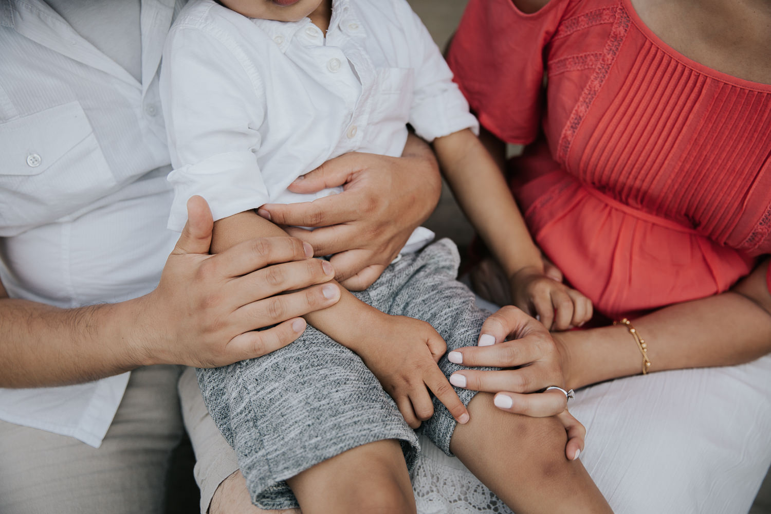 family of 3 sitting on front porch, toddler boy sitting on dad's lap with mom next to him, father and son in white shirts, mother wearing coral top, close up of hands - Barrie Lifestyle Photography