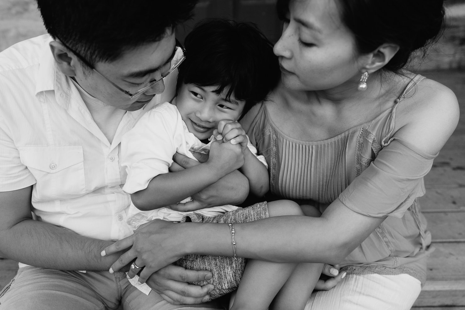 family of 3 sitting on front porch, toddler son sitting on parent's lap, snuggling into mom and dad with hands clasped as parents embrace him - GTA Lifestyle Photography