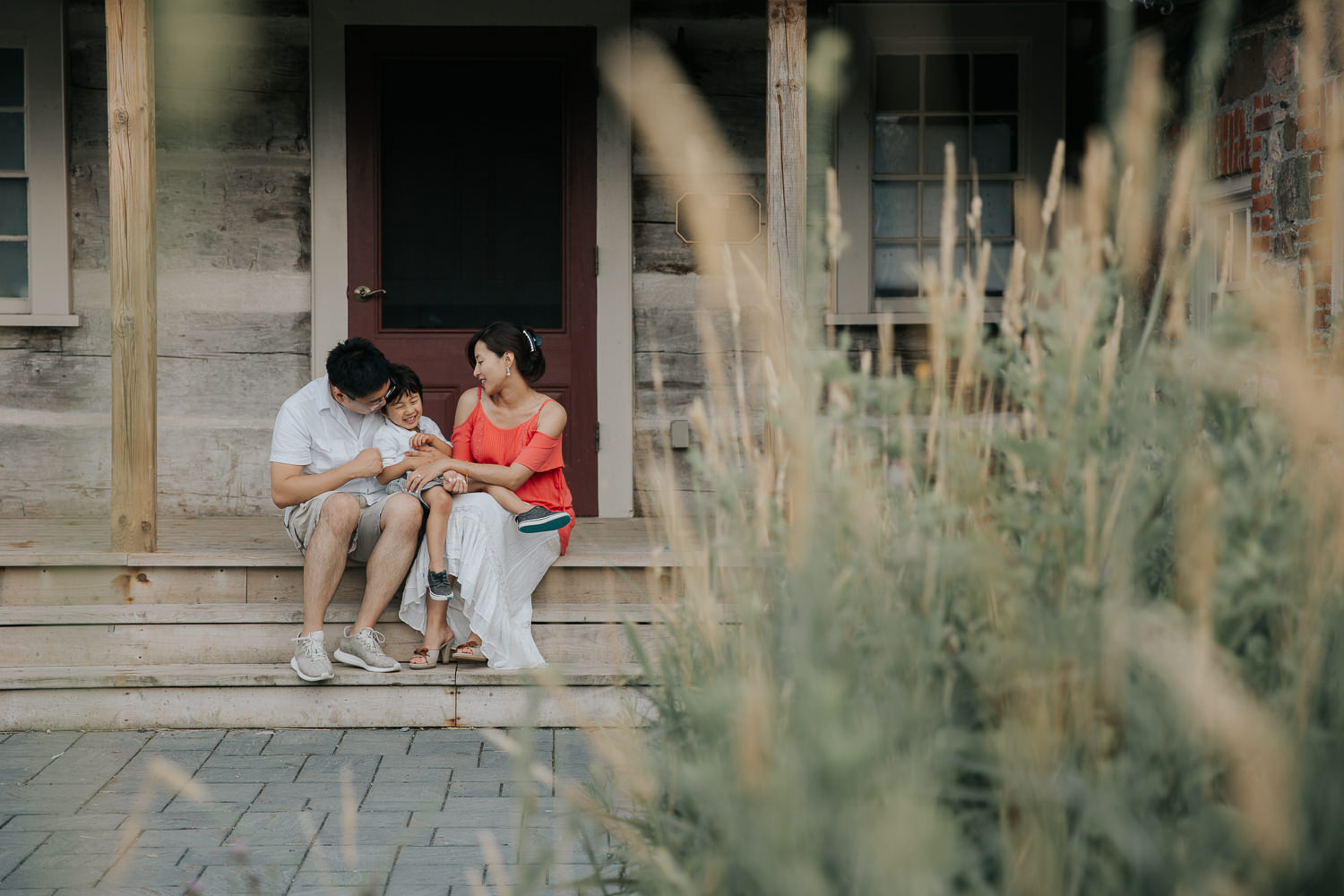 family of 3 sitting on front porch, toddler son sitting on dad's lap, laughing as mom tickles him, father and son in white shirts, mother wearing coral top - Markham Lifestyle Photography