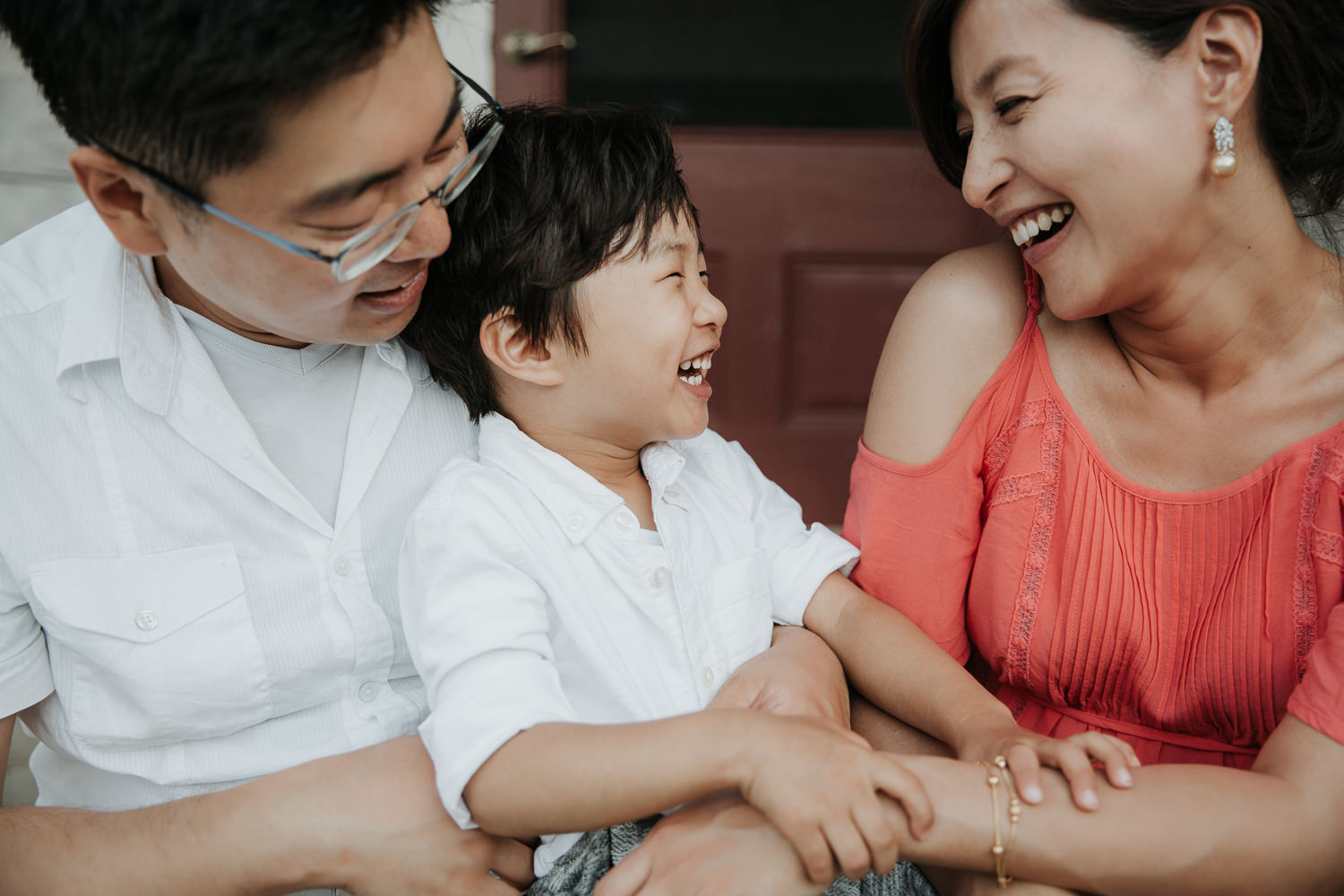 family of 3 sitting on front porch, toddler son sitting on dad's lap, looking at mom and laughing, father and son in white shirts, mother wearing coral top - Newmarket Lifestyle Photography