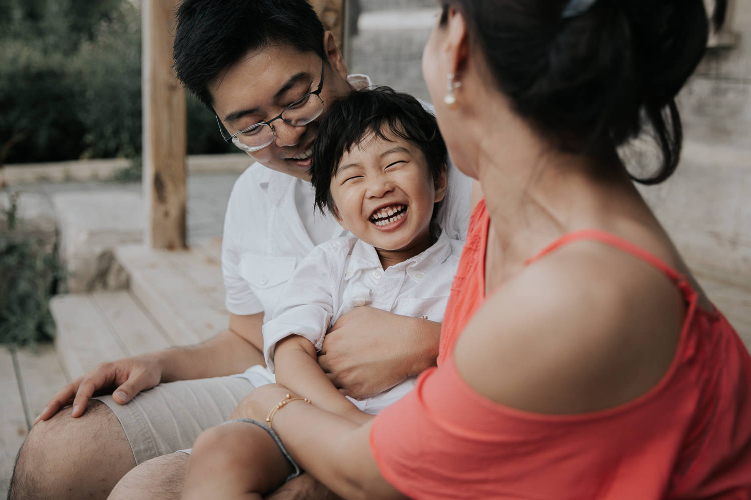 family of 3 sitting on front porch, toddler son sitting on dad's lap, laughing as mom tickles him, father and son in white shirts, mother wearing coral top - Markham In-Home Photography