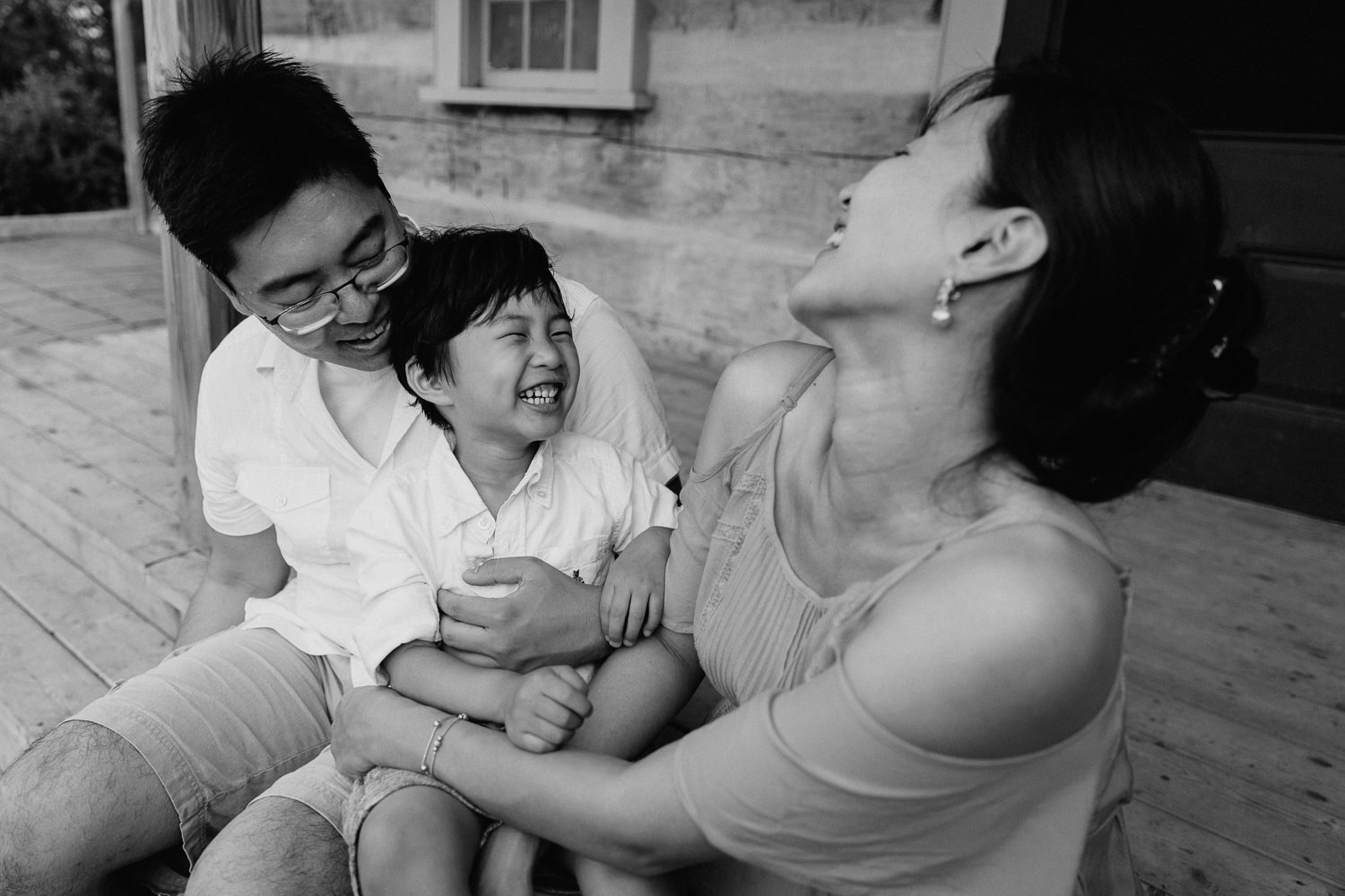 family of 3 sitting on front porch, toddler son sitting on dad's lap, laughing as mom tickles him - Stouffville In-Home Photography