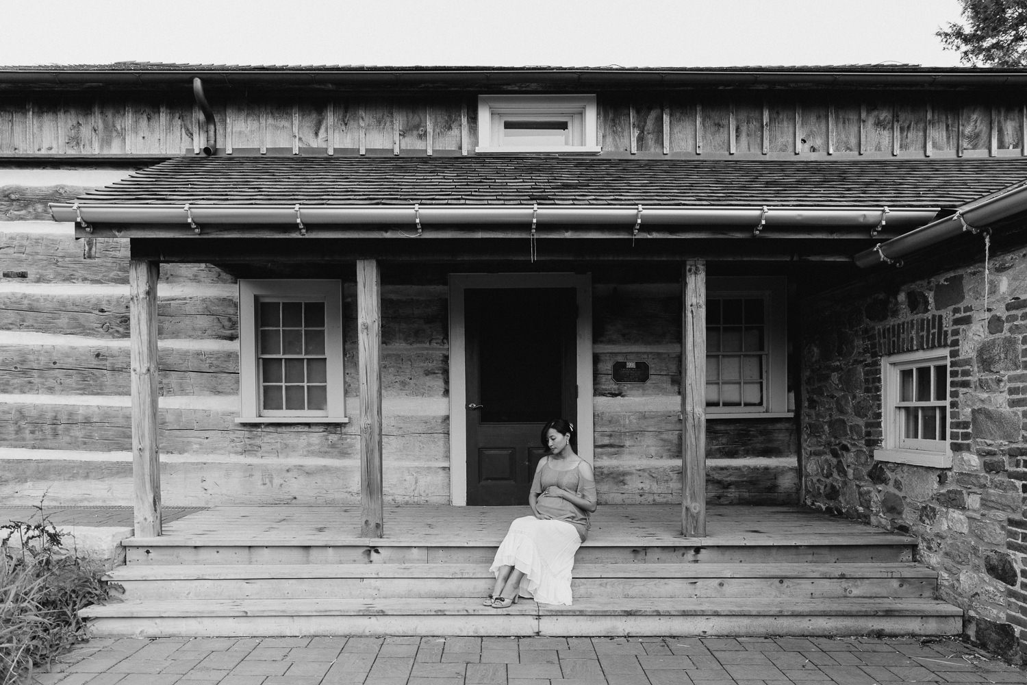 pregnant woman in flowing top and skirt sitting on porch in front of historic log home, holding baby bump - Barrie In-Home Photography