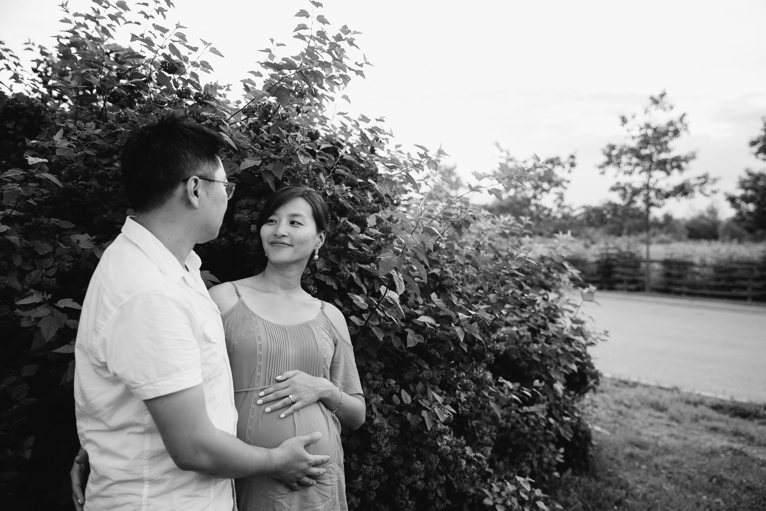 pregnant woman with brunette hair wearing coral top standing with and looking at husband, both resting hands on her baby bump - Newmarket In-Home Photos