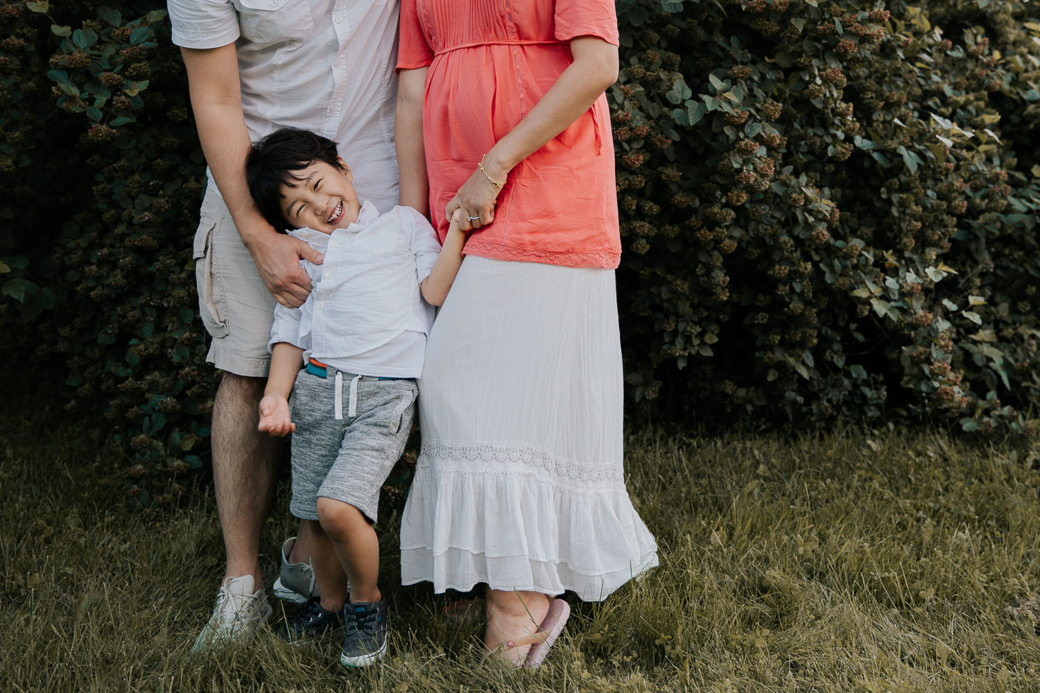 3 year old toddler boy standing in front of parents, next to mom's baby bump and holding her hand, dad's tickling him - Stouffville In-Home Photos