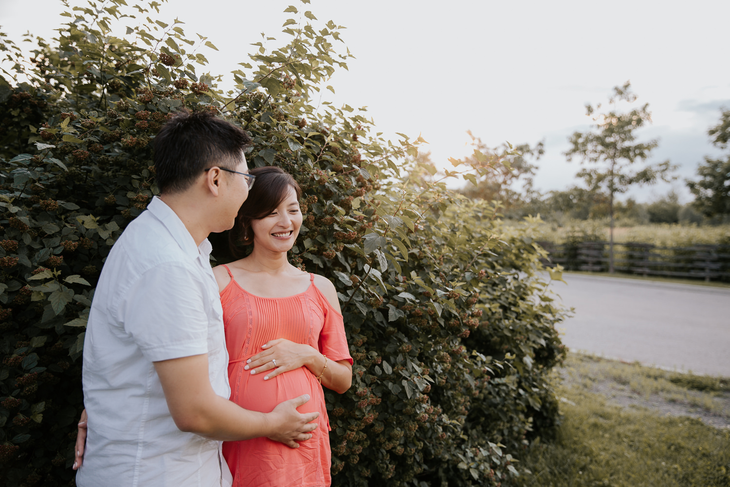 pregnant woman with brunette hair wearing coral top standing with husband, both resting hands on her baby bump - GTA In-Home Photography