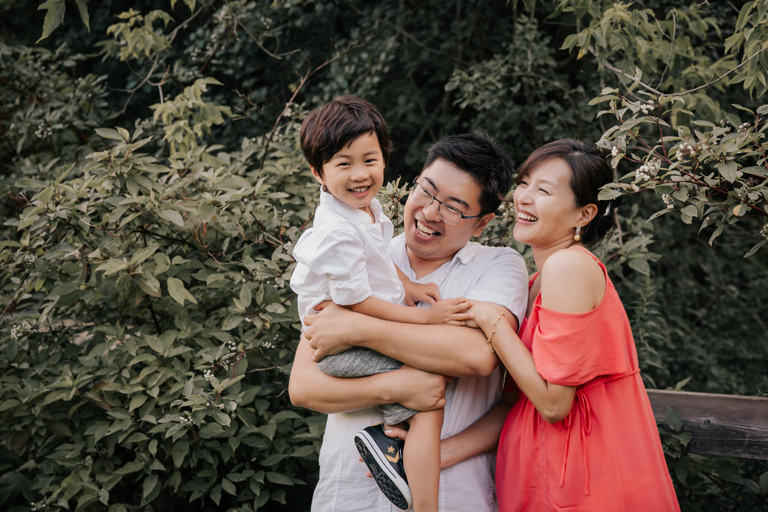 family of 3, father holding three year old toddler son who is looking at camera smiling, mom and dad looking at boy and laughing - Newmarket Lifestyle Photos