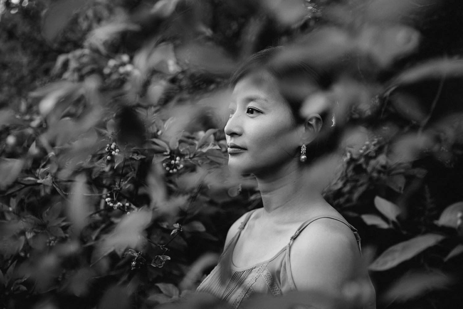 brunette woman with hair up in clip, wearing dangly earrings and a tank top looking off into the distance, shot through foliage - Markham In-Home Photos