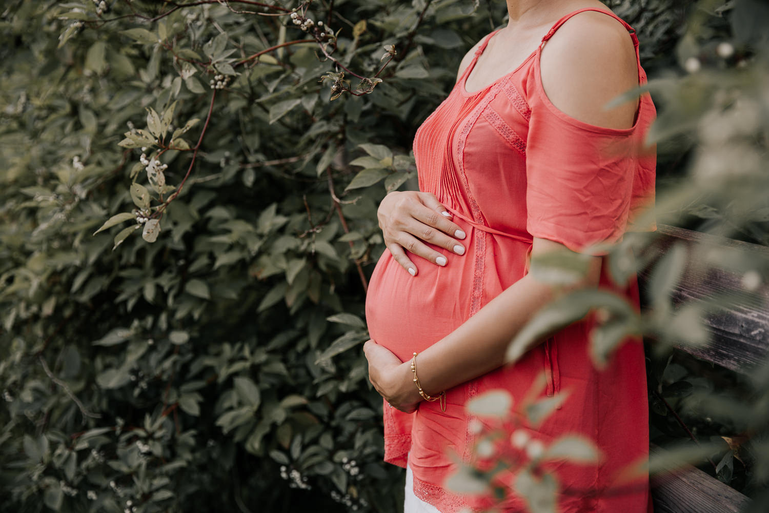 pregnant brunette woman in flowy coral top leaning against wooden fence holding baby bump - Barrie In-Home Photos