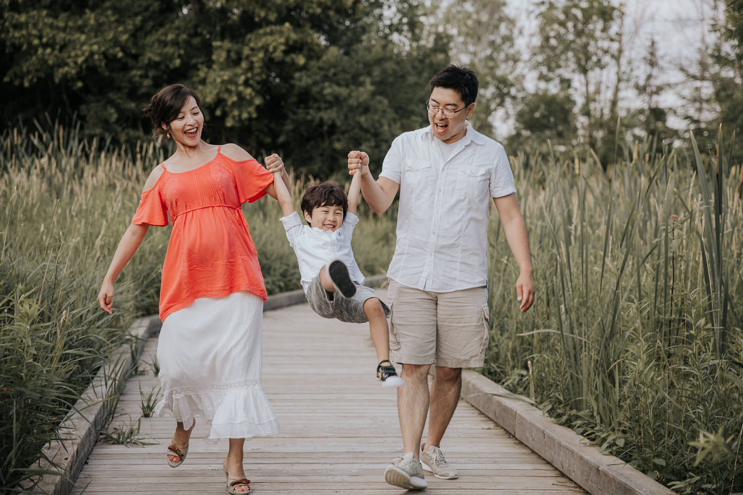 family of 3, mom and dad swing toddler boy between them as they walk down boardwalk, mom wearing flowy coral top and white skirt - Newmarket Lifestyle Photography
