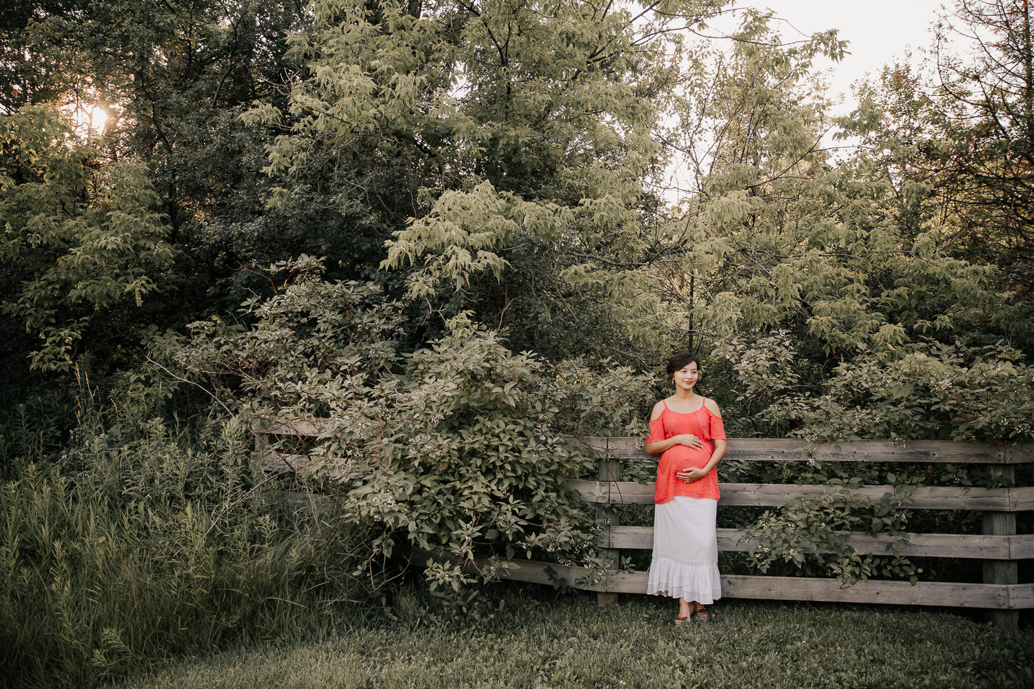 golden hour, pregnant woman in flowy coral top and white skirt leaning against wooden fence holding baby bump as sunlight filters through the trees behind her - GTA In-Home Photos