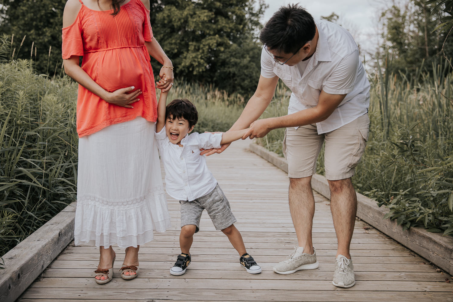 family of 3 standing on boardwalk, mom holding baby bump with one hand and toddler son's hand with other, dad tickles boy as he laughs - Stouffville Lifestyle Photography