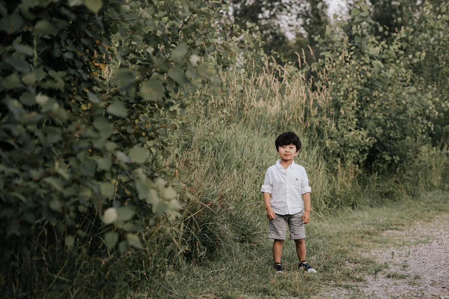3 year old boy with dark hair wearing white shirt and grey shorts standing next to long green grasses and smirking at camera -Barrie Lifestyle Photography