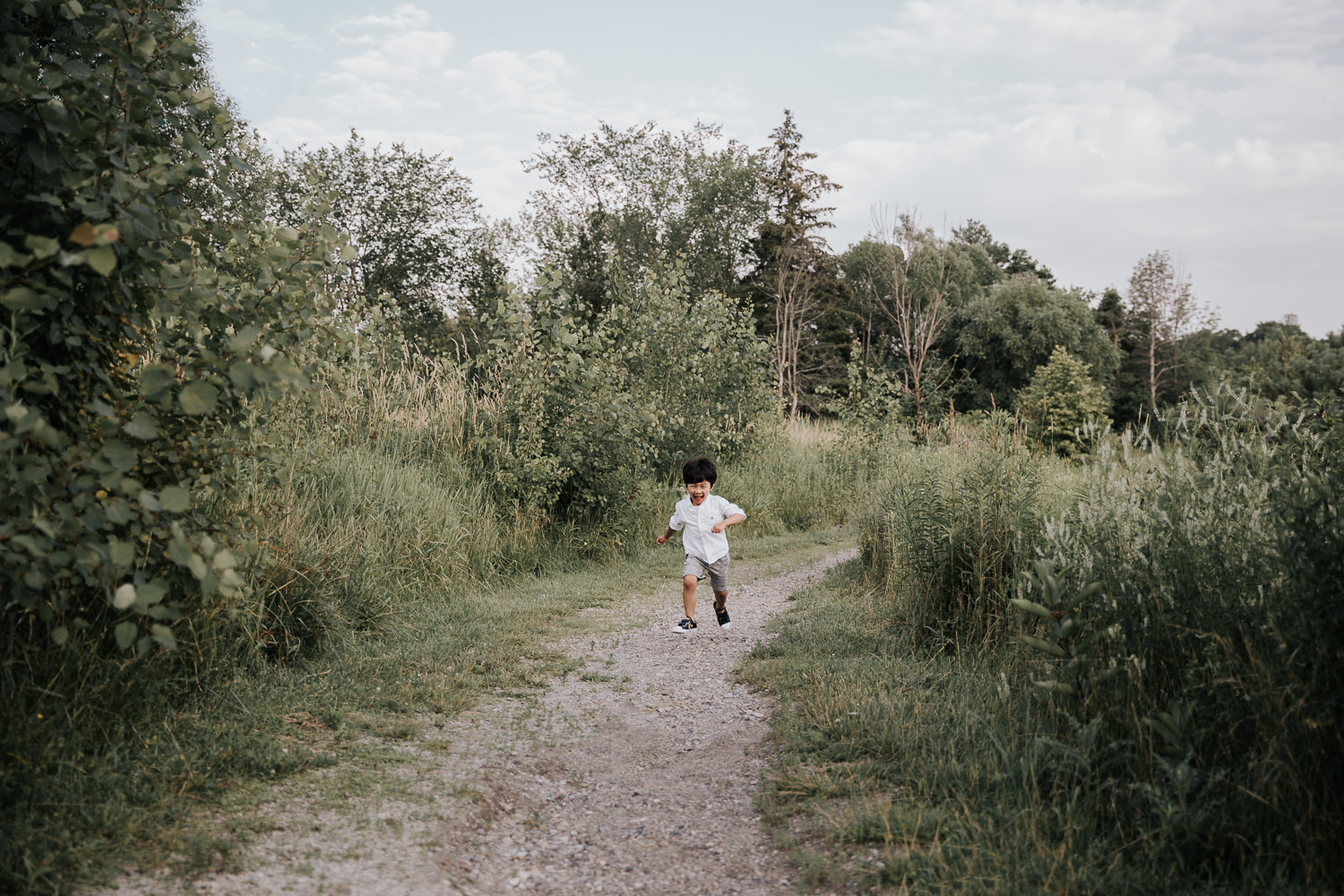 3 year old boy with dark hair wearing white shirt and grey shorts running down outdoor path smiling and full of joy -  Markham Lifestyle Photography