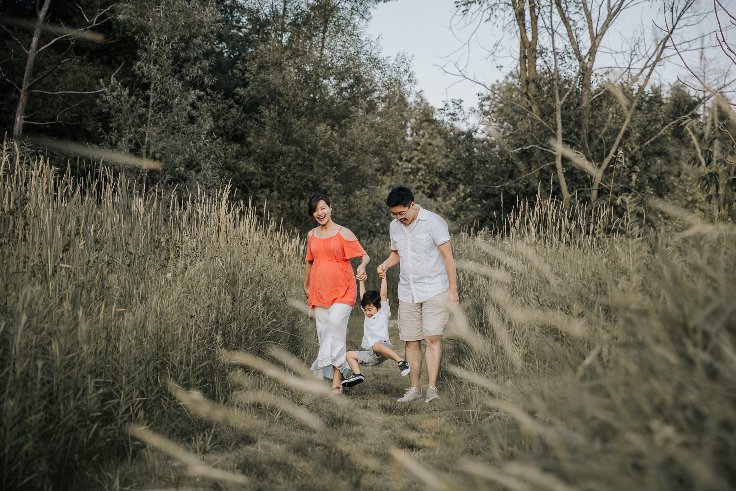 family of 3 walking down trail between long grasses and swinging toddler boy between them, everyone smiling - GTA Lifestyle Photography