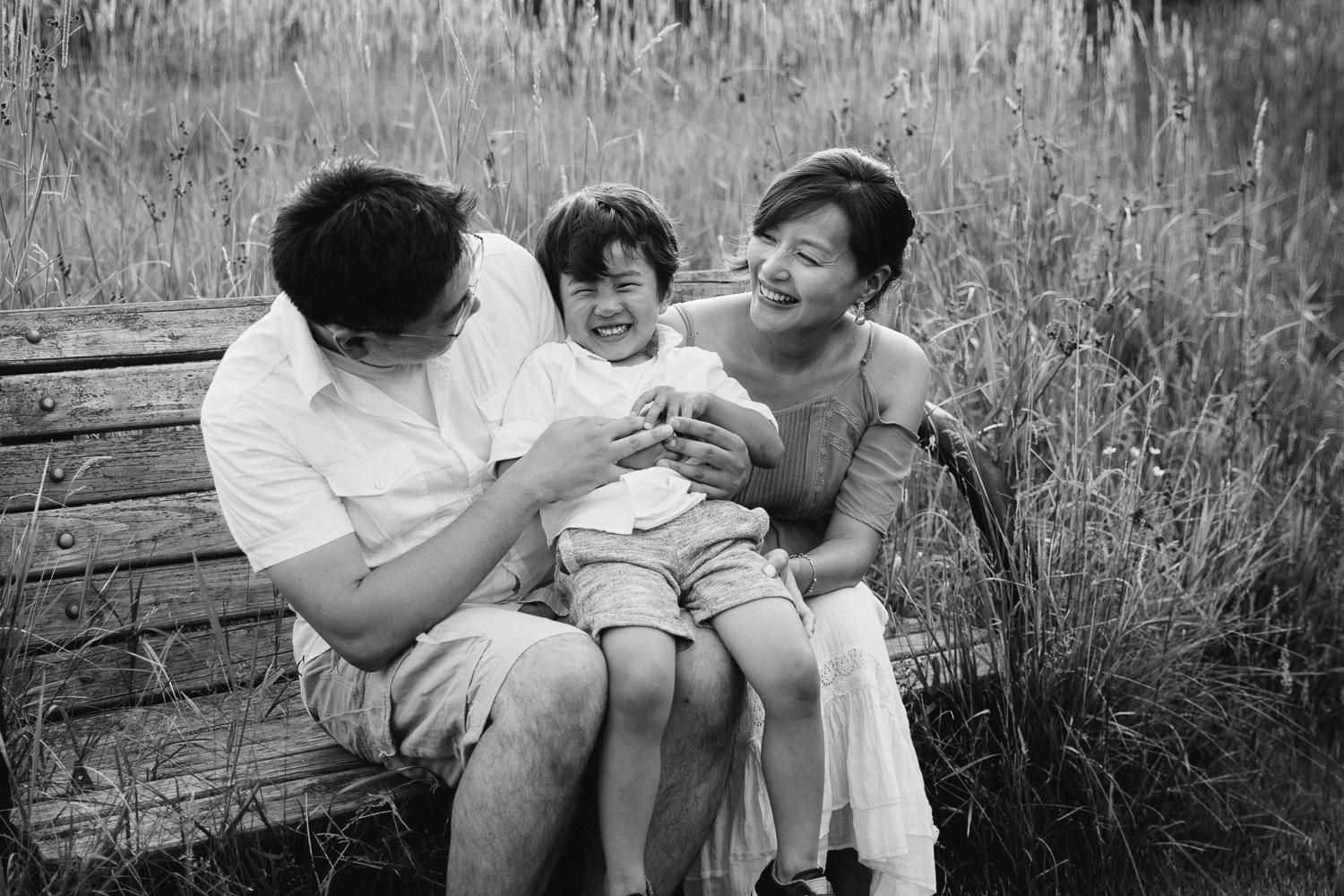 family of 3 sitting on bench park, toddler boy sitting on parent's laps and laughing as dad tickles him - Markham In-Home Photography