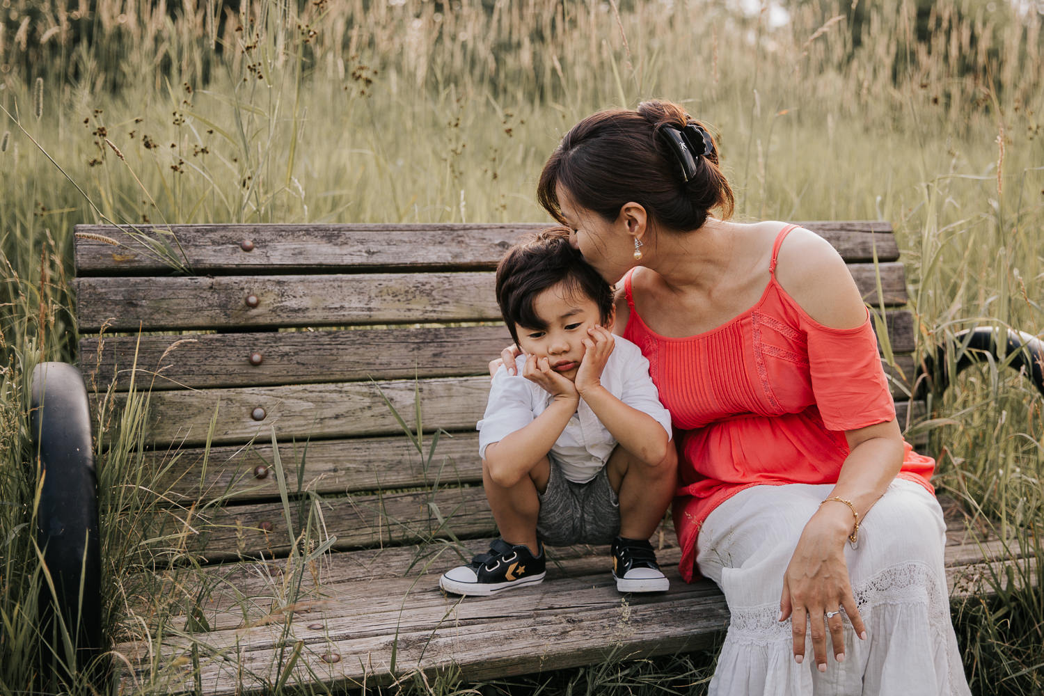 3 year old toddler boy squatting on bench with head in hands and a sad face, mom sitting next to him, leaning over and kissing son's head - Barrie In-Home Photography