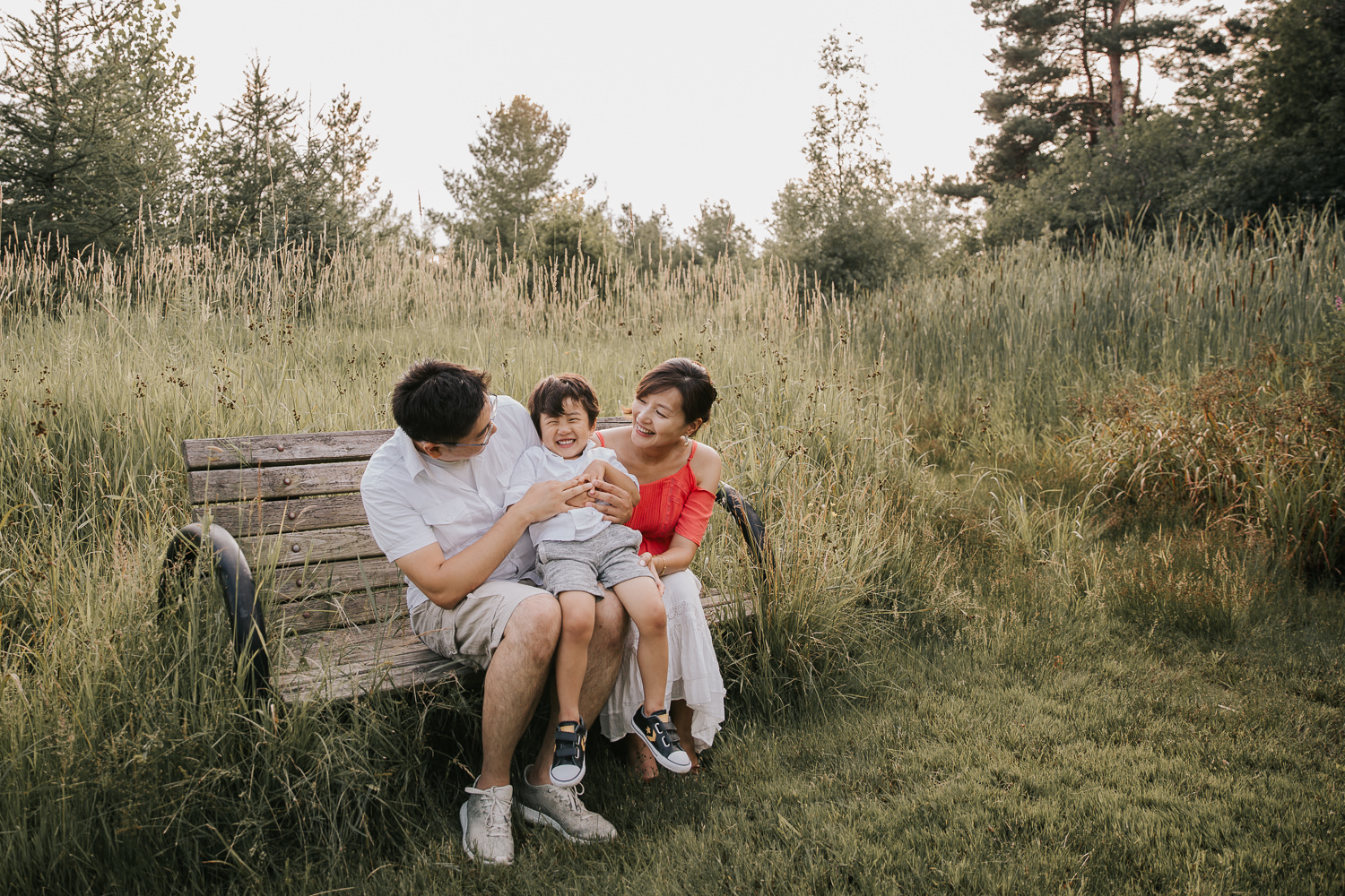 family of 3 sitting on bench park, toddler boy sitting on parent's laps and laughing as dad tickles him - Stouffville In-Home Photography