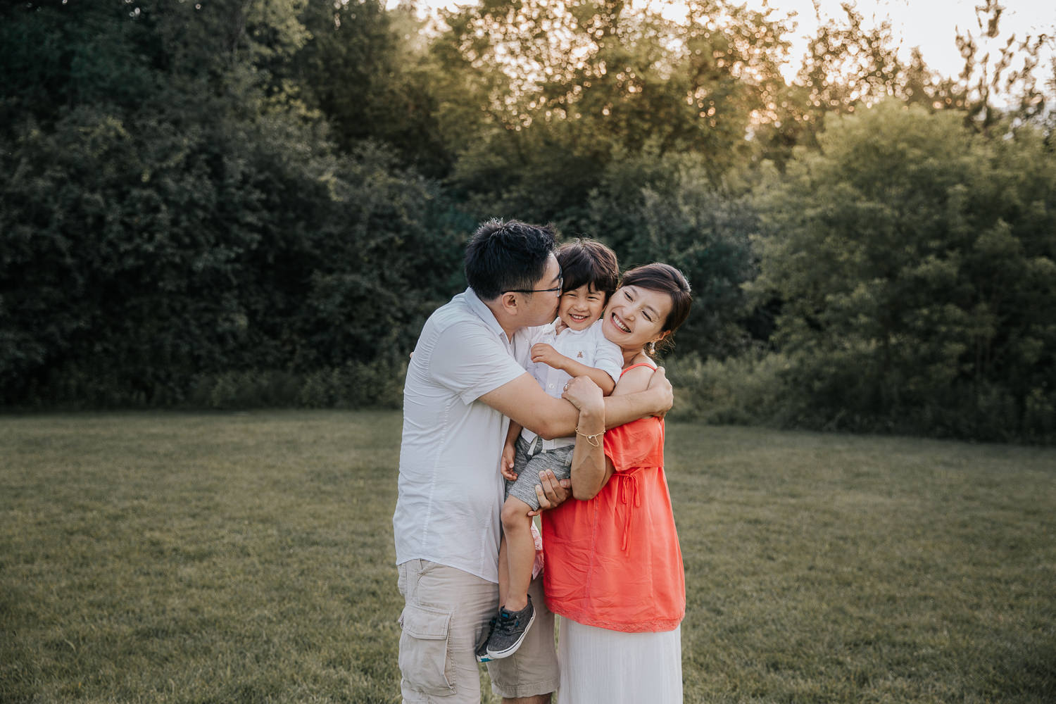 family of 3 standing in park, mom and dad embracing 3 year old toddler son between them, dad kissing boy on cheek - Markham In-Home Photos