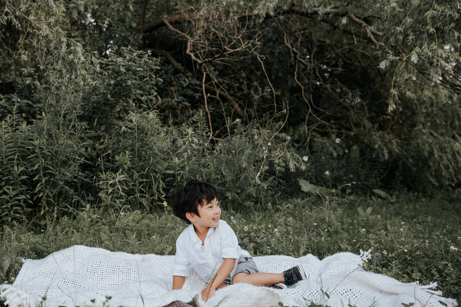  3 year old toddler boy with dark hair wearing white button down top and grey shorts sitting on white blanket in front of greenery and smiling and looking to the side - Barrie In-Home Photos