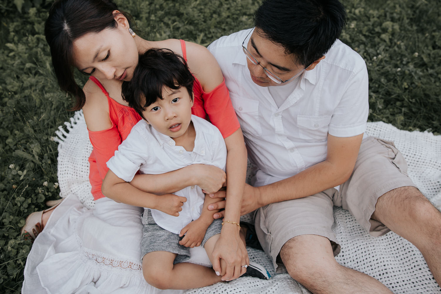 family of 3 sitting on white blanket outside, 3 year old boy sitting in mother's lap and looking up at camera, dad snuggled next to them - GTA In-Home Photos