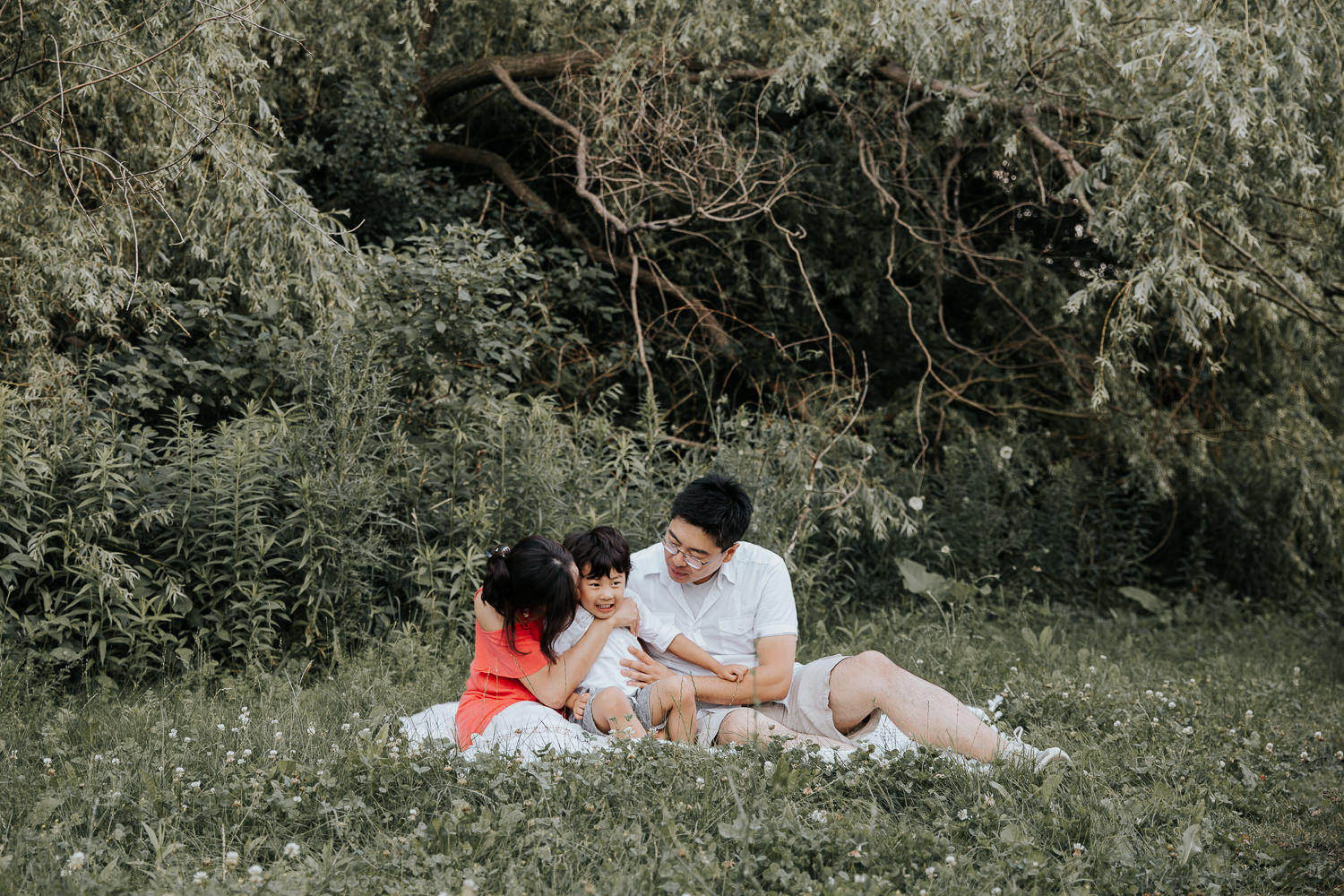 family of 3 sitting on white blanket outside, 3 year old boy sitting in mother's lap as she kisses him on cheek, dad snuggled next to them - Newmarket Lifestyle Photos