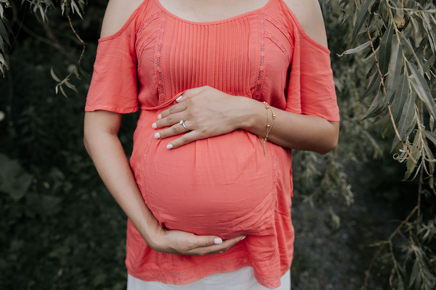 pregnant woman in flowing coral top standing in front of willow tree holding baby belly - Markham Lifestyle Photos