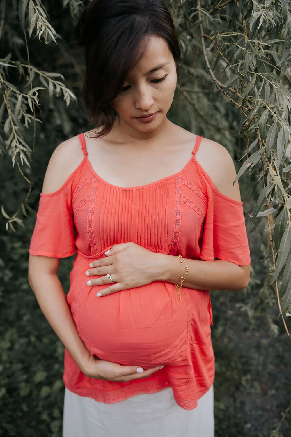 pregnant woman in flowing coral top standing in front of willow tree holding baby belly and looking down - GTA Lifestyle Photos