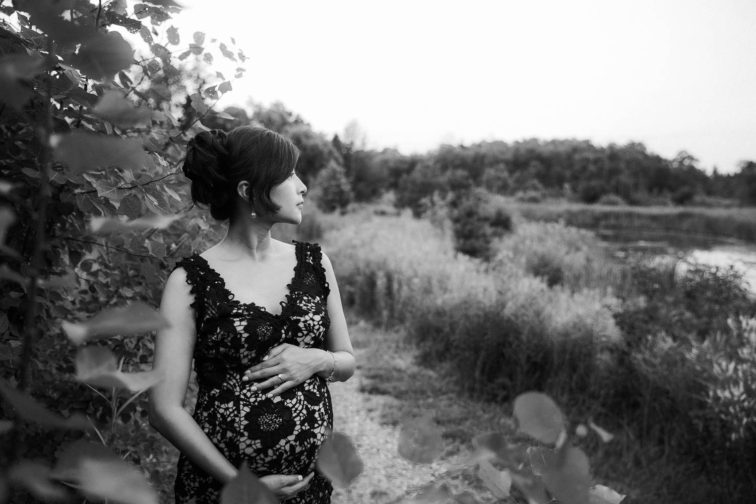 7 month pregnant woman with dark hair wearing black lace dress standing next to foliage, holding baby bump and looking off the the side - Stouffville Lifestyle Photography