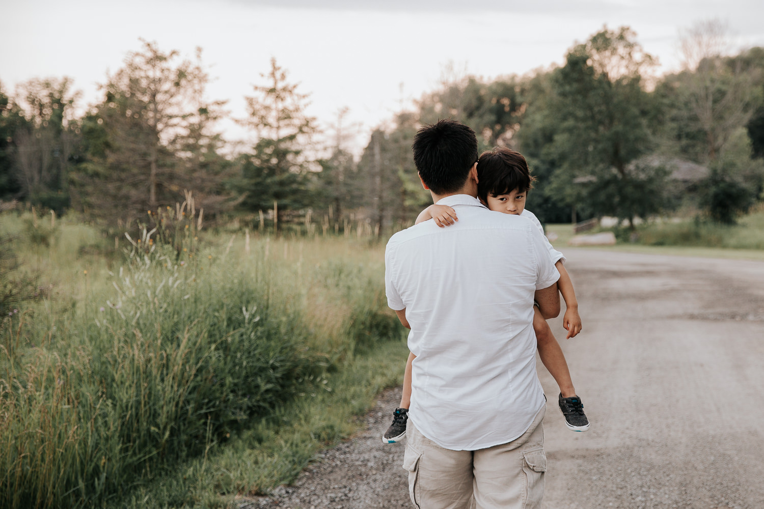 3 year old boy with dark hair looking over father's shoulder at camera as dad carries him down scenic gravel road - Newmarket Lifestyle Photography