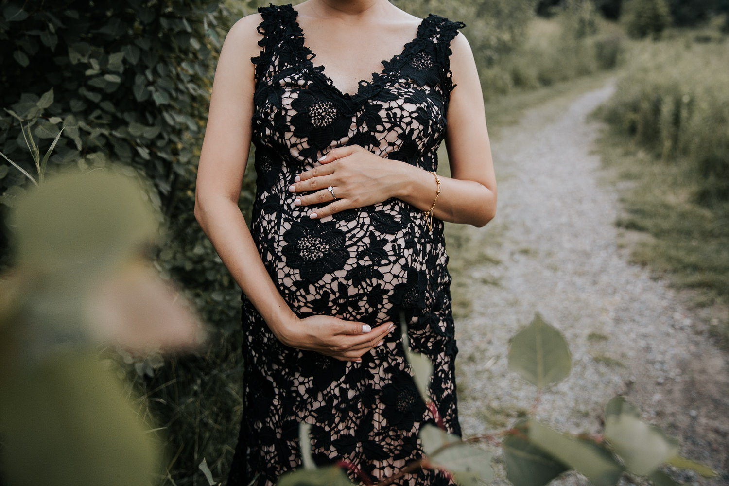 7 month pregnant woman with dark hair wearing black lace and blush gown standing next to green foliage, holding baby bump -​​​​​​​ Newmarket In-Home Photography