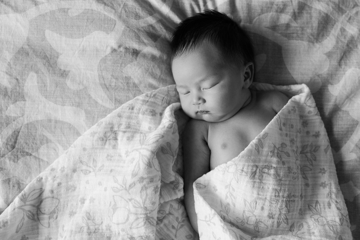 2 week old baby girl with lots of dark hair sleeping on bed half wrapped in floral swaddle and head leaning to the side - Newmarket Lifestyle Photography