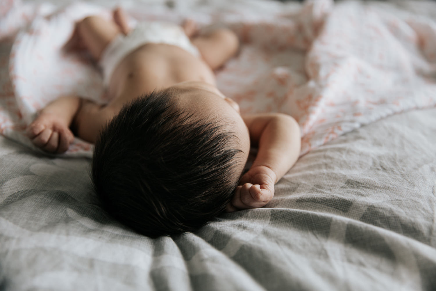 2 week old baby girl with lots of dark hair in diaper sleeping on bed on top of pink and white floral swaddle, head leaning to the side, arms in air -​​​​​​​ GTA In-Home Photography