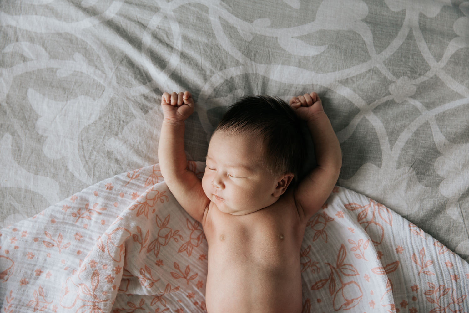 2 week old baby girl with lots of dark hair in diaper sleeping on bed on top of pink and white floral swaddle, head leaning to the side, arms in air -​​​​​​​ Markham In-Home Photography