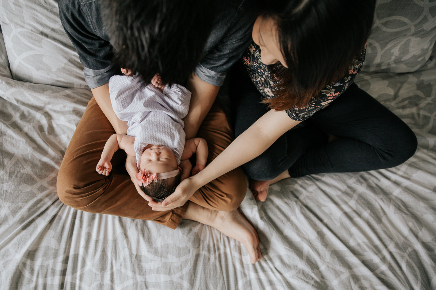 parents sitting on master bed, 2 week old baby girl with dark hair resting in dad's arms in his lap, mom's hand on daughter's head, shot from above - Stouffville In-Home Photos