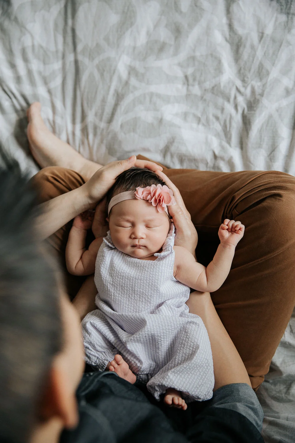 parents sitting on master bed, 2 week old baby girl with dark hair resting in dad's arms in his lap, mom's hand on daughter's head, shot of baby's face from above - Barrie In-Home Photos