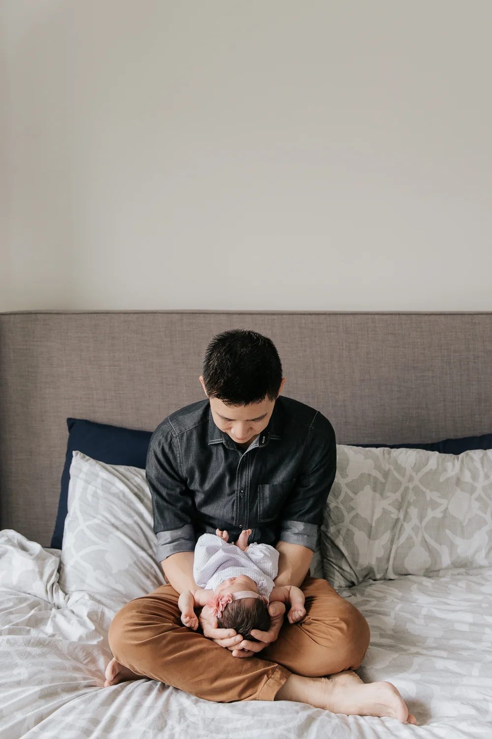 father holding and looking at 2 week old baby girl with dark hair in his arms, her head in his hands, sitting cross-legged on master bed- GTA In-Home Photos
