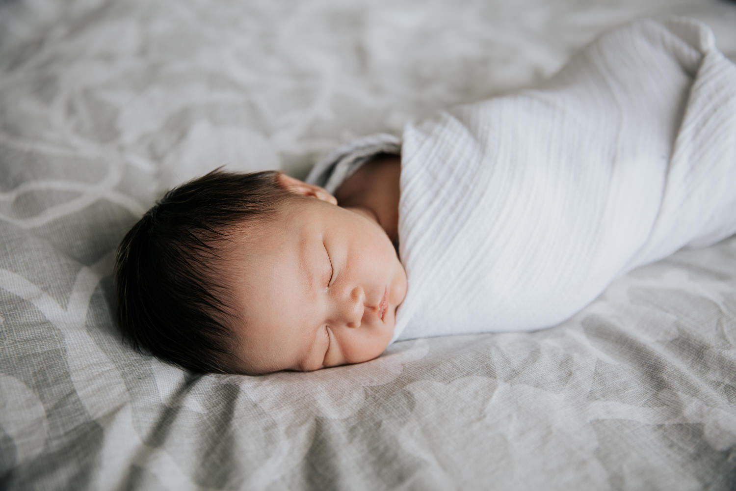 2 week old baby girl with lots of dark hair lying on bed asleep wrapped in white swaddle - Barrie Lifestyle Photography