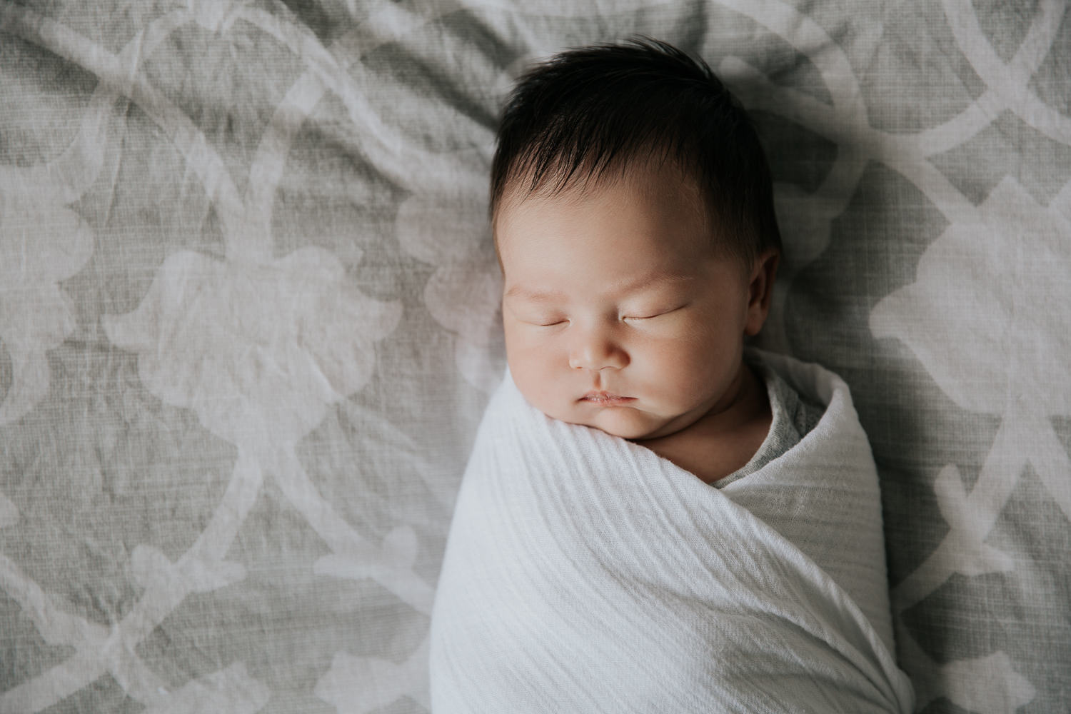 2 week old baby girl with lots of dark hair lying on bed asleep wrapped in white swaddle - GTA Lifestyle Photography