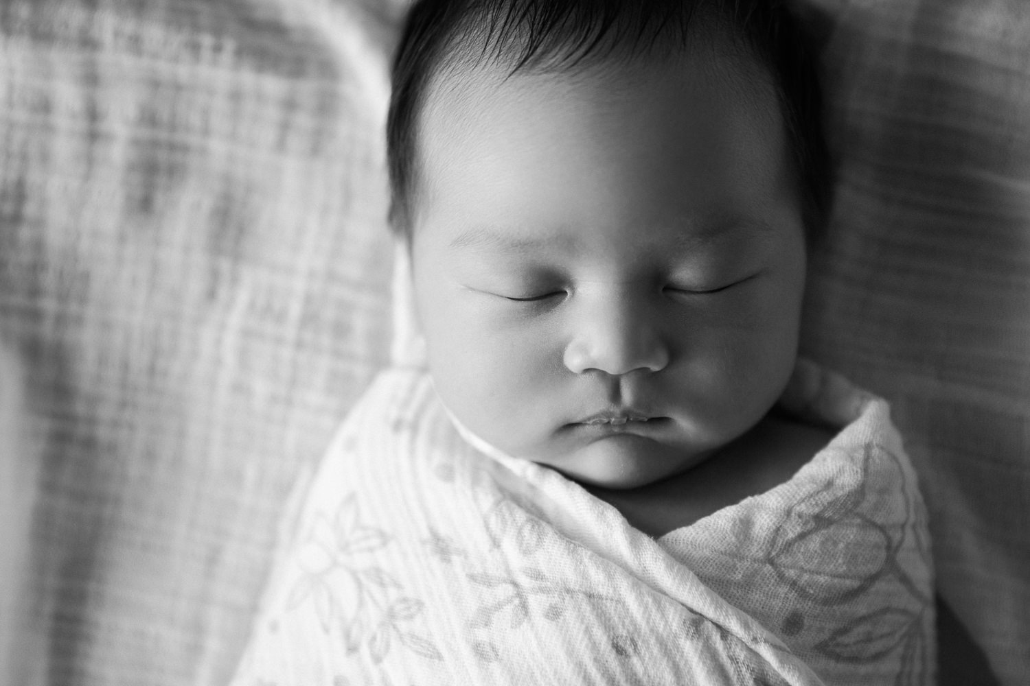 2 week old baby girl with lots of dark hair lying on bed asleep wrapped in floral swaddle, close up of face - GTA In-Home Photography