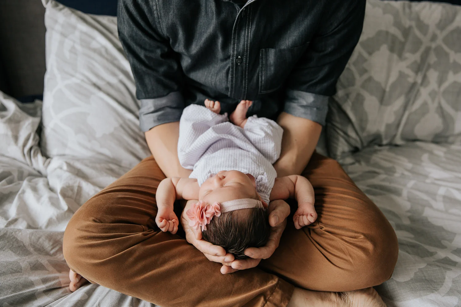 father holding 2 week old baby girl with dark hair in his arms, her head in his hands, sitting cross-legged on master bed - Newmarket Lifestyle Photos
